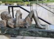 Bronze statue of a reclining person reading a book outdoors.