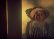 A woman in vintage attire stands by a softly lit wall, wearing a wide-brimmed hat.