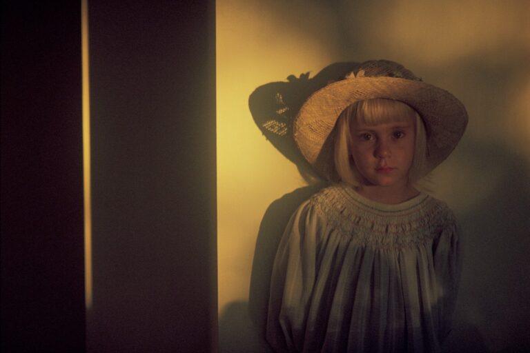 A woman in vintage attire stands by a softly lit wall, wearing a wide-brimmed hat.