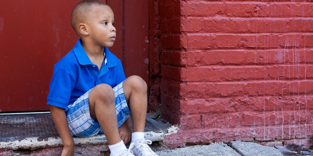 A young boy in a blue shirt sitting on the ground near a red brick wall.