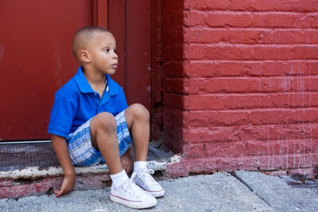 A young boy in a blue shirt sitting on the ground near a red brick wall.