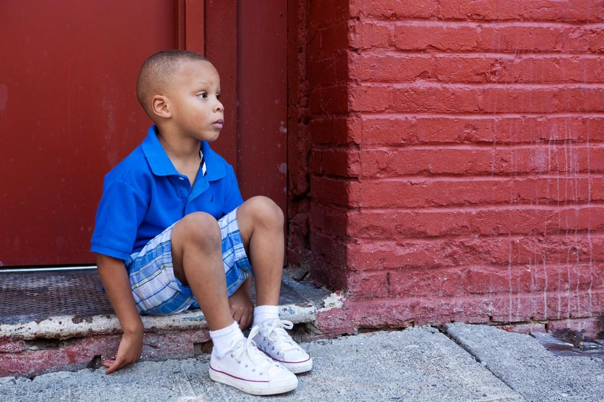 A young boy in a blue shirt sitting on the ground near a red brick wall.