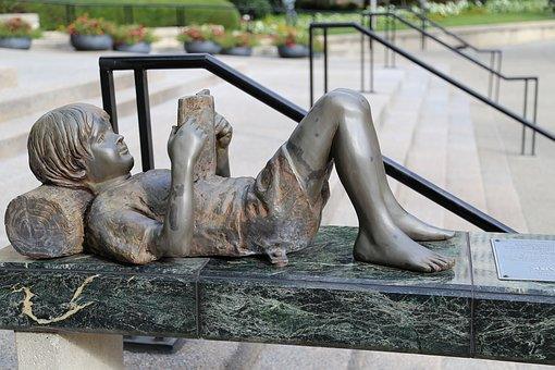 Bronze sculpture of a reclining child reading a book on a bench.
