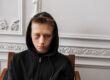Young man in black hoodie sitting against a white ornate wall.