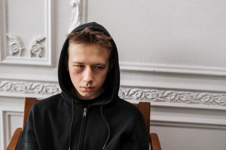 Young man in black hoodie sitting against a white ornate wall.